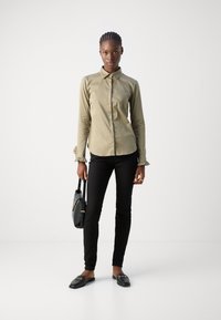 Camisa de mangas longas verde oliva com gola e punhos franzidos, combinada com calças pretas ajustadas e mocassins pretos, segurando uma pequena bolsa.