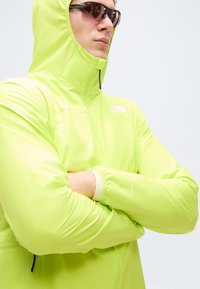 Άτομο που φοράει φωτεινό κίτρινο μπουφάν με κουκούλα και φερμουάρ κλειστό μέχρι τον γιακά, και σκούρα γυαλιά ηλίου, με σταυρωμένα χέρια μπροστά σε απλό φόντο.