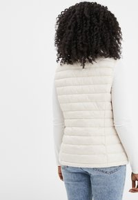Hellbeige Steppweste mit horizontalen Nähten, hohem Kragen und einem glatten, glänzenden Material; kombiniert mit einem weißen Langarmoberteil und Denim.