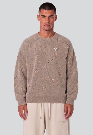 Mand iført en taupe tekstureret sweater med små huller og tekstmønstre, kombineret med lyse beige snørebukser.