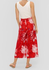 Femme portant un haut sans manches blanc associé à un pantalon large à imprimé floral rouge et des sandales beiges, se tenant devant un fond uni.