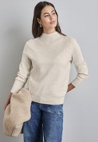 Sweter w kolorze kremowym z prążkowanym wysokim dekoltem i wzorem w zygzak na klatce piersiowej, uzupełniony podwiniętymi rękawami i fakturowanym materiałem.