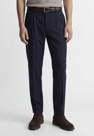 Homme portant un pantalon slim bleu marine, une ceinture en cuir marron et des bottines en daim marron foncé, debout devant un fond uni.