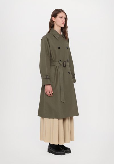 Olívazöld trench coat, dupla soros gombos, övvel ellátott derék, és mandzsettagombokkal. Bepleten egy bézs rakott szoknyával és fekete bokacsizmával.