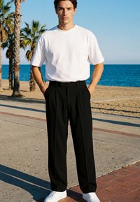 Ein junger Mann steht auf einem Strandweg, trägt ein weißes T-Shirt, schwarze weite Hosen und weiße Sneakers, mit Palmen und Ozean im Hintergrund.