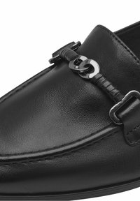 Gros plan sur une chaussure bateau en cuir noir avec une chaîne en métal argenté et un détail de sangle en cuir sur le dessus.