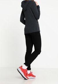 Persoon in zwarte broek en een donkergrijze hoodie, staand met één been gekruist, draagt felrode sneakers en witte Adidas sokken.