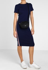 Marineblauwe jurk tot de knie met korte mouwen, voorzien van rode en witte zijstrepen; gecombineerd met een zwarte schoudertas en witte sneakers.