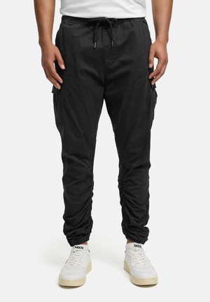 Mężczyzna w czarnych joggerach typu cargo z elastycznymi mankietami i białych tenisówkach na jednolitym jasnym tle.