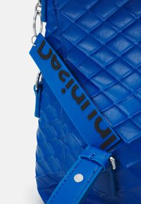Blaue gesteppte Tasche mit strukturierter Oberfläche, ausgestattet mit einem schwarzen Logo-Tag und einem passenden Trageriemen. Enthält silberne Hardware-Akzente.