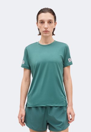 En ung person med kort, tilbagelagt hår iført en turkis atletisk kortærmet trøje og matchende shorts, stående mod en ensfarvet baggrund.