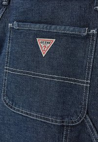Poche en tissu denim avec des coutures blanches et une petite étiquette triangulaire rouge et blanche du logo Guess Originals centrée sur la poche.