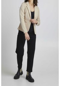 Beige sáčko na míru s klopovým límcem, kombinované s černým topem, černými kalhotami a černými kotníkovými botami s lehkými akcenty.