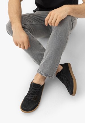 Osoba sediaca s prekríženými nohami, oblečená v sivých rifliach, čiernych semišových teniskách s gumovou podrážkou a čiernom tričku, na bielom pozadí.