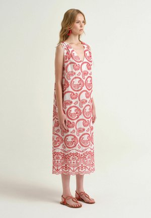 Donna che indossa un vestito midi bianco senza maniche con motivi paisley rossi, orecchini rossi e sandali rossi abbinati, in piedi di profilo su uno sfondo semplice.