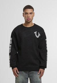 Black knitted sweater with a crew neck, featuring gray logo on chest and "BRAND" text along the sleeves. Made from soft, textured fabric.