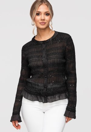 Femme blonde portant un chemisier noir transparent boutonné avec des volants et des sequins, associé à un pantalon blanc, posant devant un fond uni.