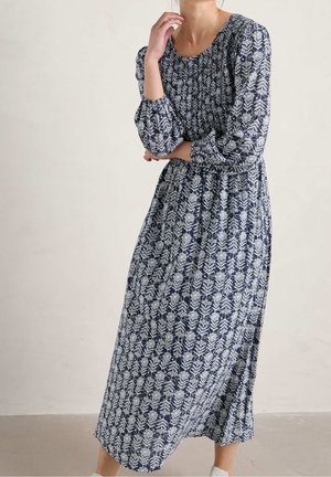 Frau in einem marineblauen Midi-Kleid mit weißem Blumenmuster, langen Ärmeln, steht mit einem verschränkten Arm und der anderen Hand nahe ihrem Gesicht.