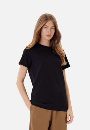 Donna con lunghi capelli biondi che indossa una maglietta nera a maniche corte e pantaloni marroni, in piedi con una mano in tasca davanti a uno sfondo semplice.