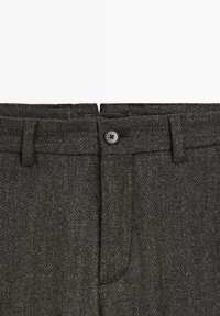 I pantaloni grigi a spina di pesce presentano una texture liscia, una cinta sartoriale e una chiusura frontale con bottone nero. Due tasche frontali incluse.