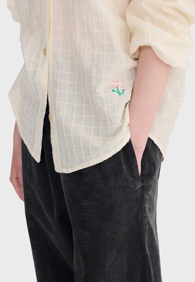 Personne portant une chemise à carreaux crème à boutons, brodée d'une petite fleur rose et verte, et un pantalon noir, une main dans la poche.