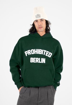Mand iført en mørkegrøn hættetrøje med "PROHIBITED BERLIN" tekst, grå bukser og en hvid strikket hue med en mærkat, står med hænderne i lommerne.