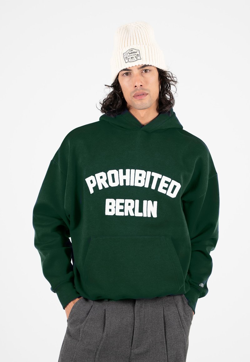 Homme portant un sweat à capuche vert foncé avec le texte "PROHIBITED BERLIN", un pantalon gris et un bonnet en tricot blanc avec un écusson, debout les mains dans les poches.