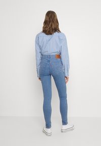 Jeans skinny de talle alto de mezclilla azul con un parche de cuero de la marca; combinados con una camisa de botones a rayas en azul y blanco y zapatillas blancas.