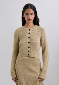 Woman with long dark hair wearing a beige textured knit cardigan with black buttons and matching skirt, standing against a plain background.
