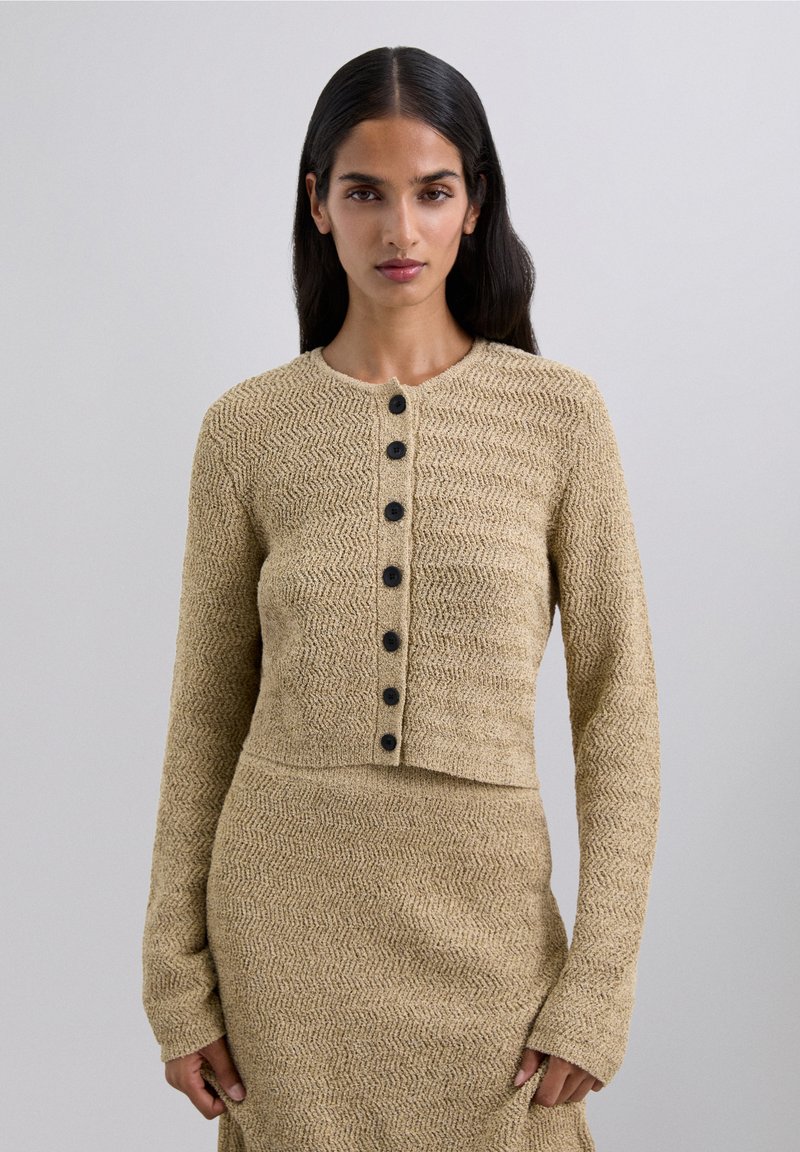 Woman with long dark hair wearing a beige textured knit cardigan with black buttons and matching skirt, standing against a plain background.