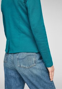 Haut à manches longues couleur sarcelle avec une finition texturée et un ourlet arrondi, associé à un jean taille haute bleu clair avec des poches arrière.