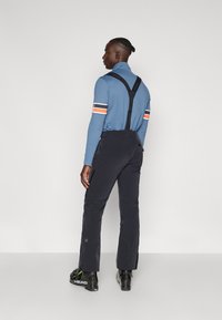 Blaues Langarmhemd mit weißen und orangefarbenen Streifen sowie schwarzen Skihosen mit Trägern. Schwarze Skischuhe mit grünen Akzenten.