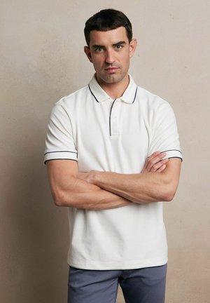 Biała koszulka polo z kołnierzykiem, z granatowym wykończeniem na rękawach i kołnierzyku; krótki rękaw i luźny fason. W zestawie z szarymi spodniami.