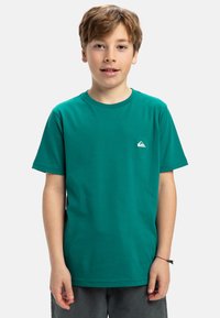 Zelené tričko s okrúhlym výstrihom vyrobené z bavlny, s krátkymi rukávmi a malým bielym logom na ľavej hrudi. Hladká textúra, voľný strih.