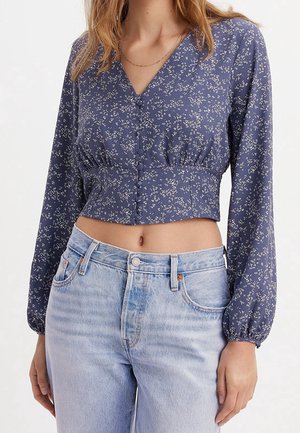 Femme portant un chemisier court à manches longues bleu à motif floral avec des boutons recouverts de tissu et un jean taille haute bleu clair.