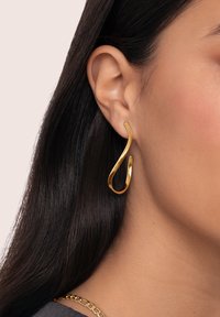 Boucle d'oreille en or avec un design ondulé, présentant une surface polie et réfléchissante, accrochée au lobe de l'oreille, complétant des cheveux droits et foncés.