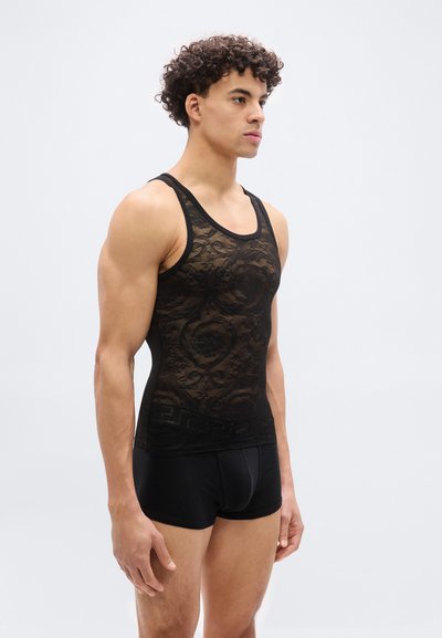 Άνδρας που στέκεται, φορώντας μαύρο διάφανο δαντελένιο τοπ χωρίς μανίκια και μαύρα εφαρμοστά μποξέρ, μπροστά σε απλό ανοικτό γκρι φόντο.