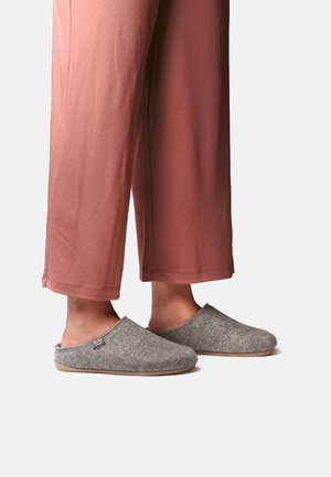 Person iført løse lyserøde bukser og grå slip-on filt tøfler med flade såler på en ensfarvet baggrund.