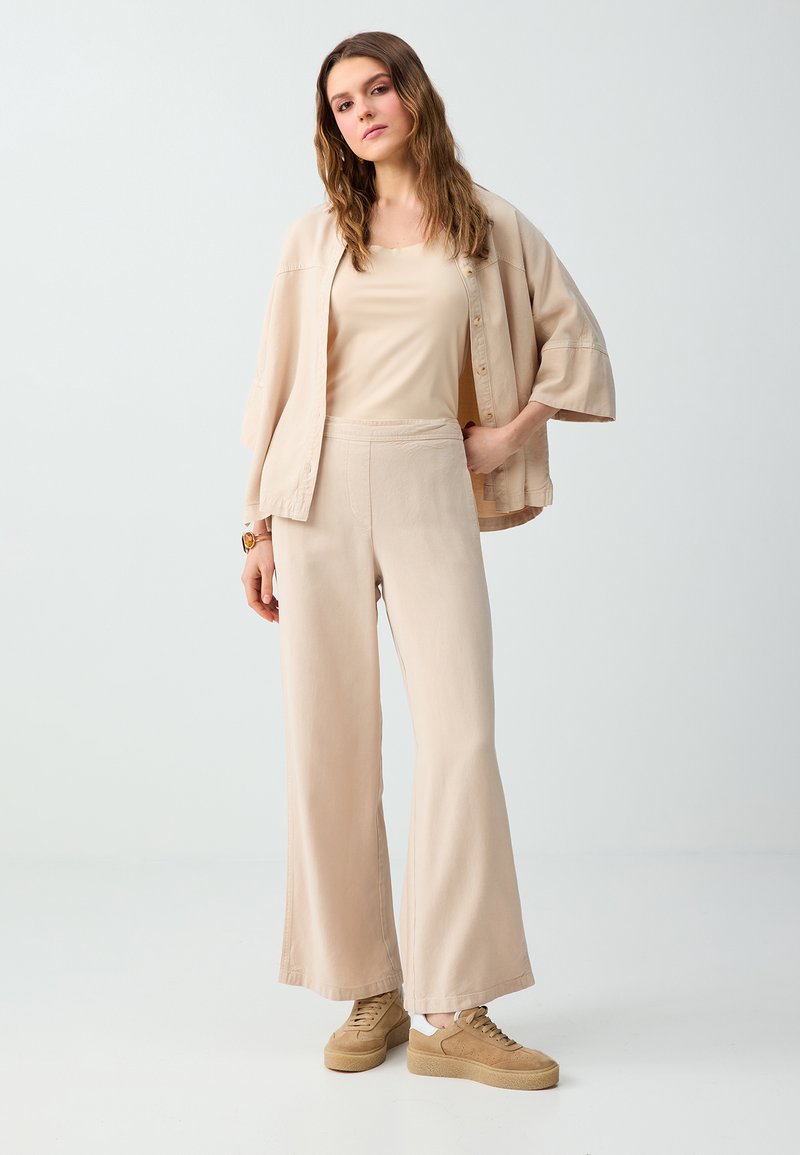 Femme aux longs cheveux bruns portant un pantalon beige à jambes larges, un haut assorti, une veste ample et des baskets compensées couleur beige, debout devant un fond clair uni.