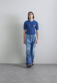 Blaues Netz-Polohemd mit Kragen, kombiniert mit hellen, ausgestellten Jeans. Das Modell steht vor einem neutralen, strukturierten Hintergrund.