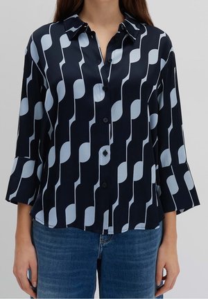 Femme portant une chemise noire à boutons avec un motif géométrique vertical bleu clair et un jean bleu, debout devant un fond uni.