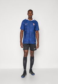 Tricou de fotbal albastru cu un model texturat, guler alb și emblema Franței. Șorturi și șosete negre cu brandul Nike; ghete negre.