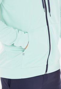 Lys grøn zip-jakke med en glat tekstur, der har en frontlomme og mørkeblå detaljer på lynlåsen og ærmeafslutningerne.