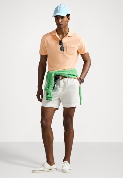 Lichtoranje polo shirt, witte shorts, groene trui vastgebonden om de taille en blauwe pet; casual schoeisel; strakke, minimalistische stijl.