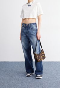 Avklippt vit t-shirt med marinblå logga, blå högmidjade jeans, svart-vita sneakers och en liten brun tigerrandig handväska.