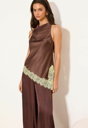 Naine, kes kannab varrukateta pruuni satiintoppi kahvatu kollase pitsiäärega ja sobivaid laia säärega pükse, seisab lihtsa heleda tausta ees.