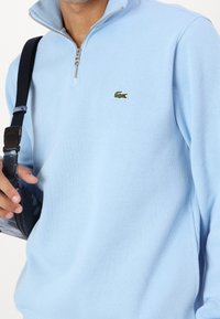 Ljust blå texturerad halvdraget tröja med ribbat mönster och en liten grön krokodil-logotyp på vänster bröst.