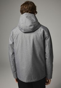 Jachetă gri cu glugă, dintr-un material texturat, cu o croială relaxată, cusături pronunțate și un tiv curbat. Design simplu, fără logo-uri sau embellishments vizibile.