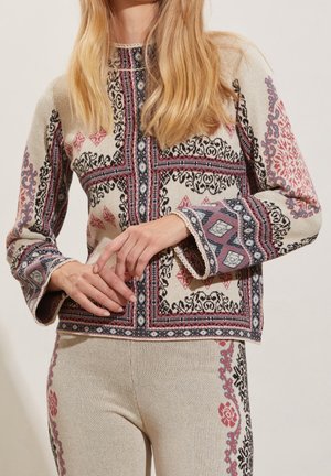 Strikket beige sweater og bukser med indviklede sorte, lilla og pink blomster- og geometriske mønstre, båret af en person med langt blondt hår.