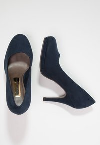 Un par de zapatos de tacón alto de ante azul marino, uno visto desde arriba y el otro de lado, sobre un fondo blanco.